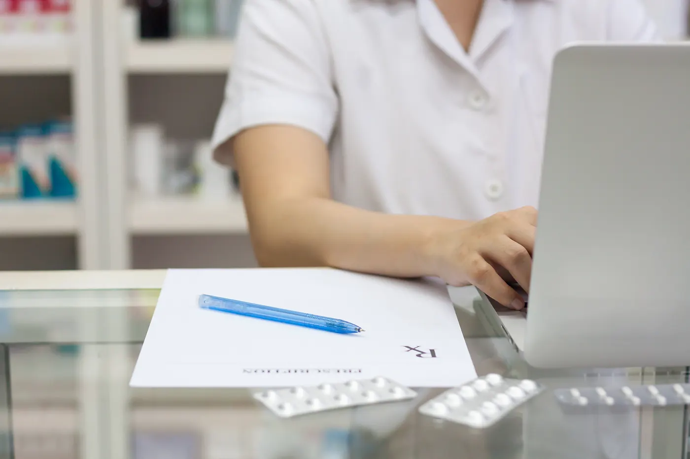 Pharmacist using a laptop at the counter with a prescription form and blister packs of tablets